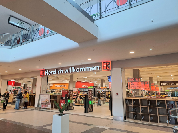 Innenansicht eines Einkaufszentrums mit Menschen, Geschäften, Infotafeln mit der Aufschrift "Herzlich willkommen", Beleuchtung, Geländern und einem Topf mit Pflanze am Boden.