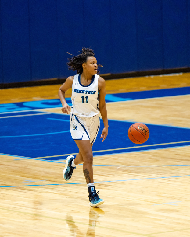 Eine Frau in einer blauen und weißen Uniform, die einen Basketball auf einem Court dribbelt, trägt ein weißes T-Shirt mit der Aufschrift "Wake Tech Women's Basketball" und blaue Schuhe, vor einer blauen Wand.