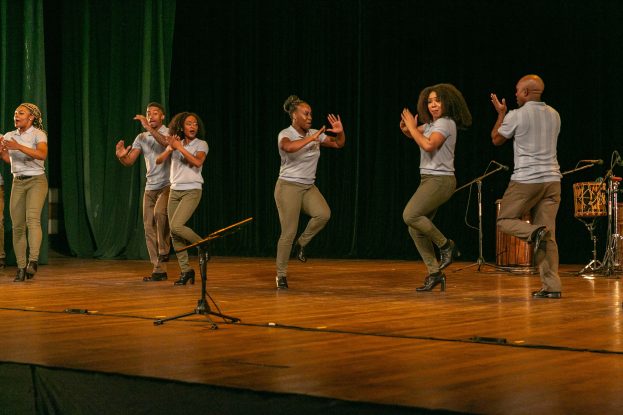 Gruppe von Menschen auf einer Bühne, die jubeln und mit Mikrofonen, Musikinstrumenten und einem Vorhang im Hintergrund.