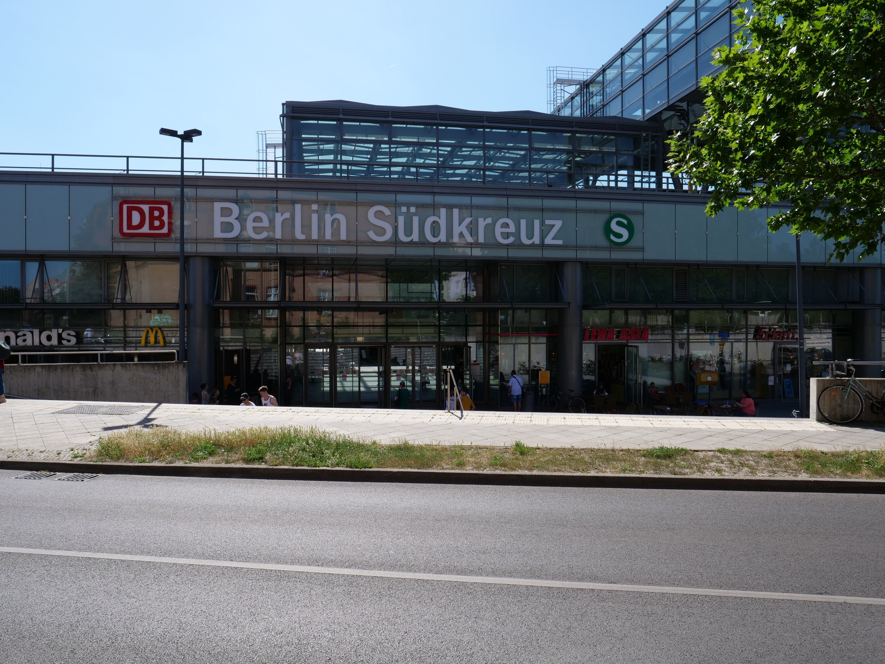 Große Glasfassade des Berliner Südkreuz-Bahnhofs mit Text an der Fassade, umgeben von Straßenmasten, Lichtern, Fahrzeugen, Füpßen, Fahrrädern, Bäumen und einem klaren blauen Himmel.