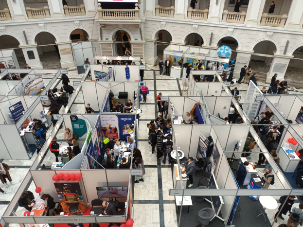 Große Besuchergruppe auf einer internationalen Fachmesse mit Ständen, Bannern, Tischen, Stühlen und Ballons in einem Raum mit architektonischen Bögen und Säulen.