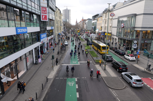 Eine belebte Stadtstraße mit Fahrradfahrern und Fahrzeugen, gesäumt von Gebäuden, Laternen und Schildern, unter einem klaren blauen Himmel mit Bäumen im Hintergrund.