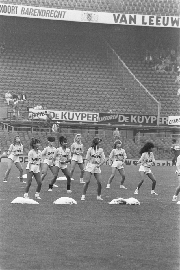Schwarz-weißes Foto einer Gruppe von Frauen auf einem Fußballfeld mit Zuschauern auf den Rängen und Bannern im Hintergrund.