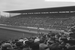Schwarzes und weißes Foto eines vollen Stadions mit Zuschauern, die ein Fußballspiel verfolgen, einige stehen und andere sitzen auf den Tribünen, mit Bannern, Pfählen, einem Schuppen, Bäumen, einem Turm und einem bewölkten Himmel im Hintergrund.