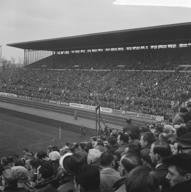 Schwarzes und weißes Foto eines vollen Stadions mit Zuschauern, die ein Fußballspiel verfolgen, einige stehen und andere sitzen auf den Tribünen, mit Bannern, Pfählen, einem Schuppen, Bäumen, einem Turm und einem bewölkten Himmel im Hintergrund.