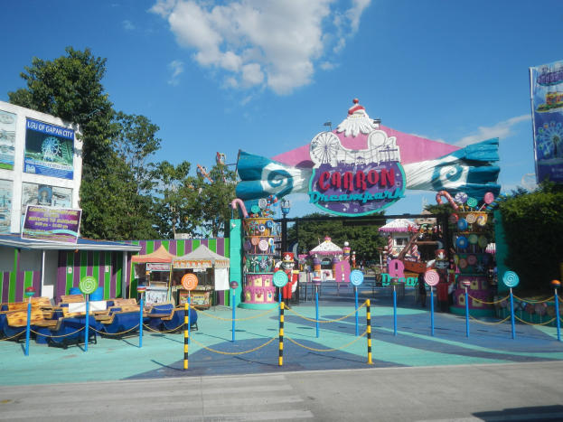 Karussell am Parkeingang mit Straße, Umzäunung, Buden, Schildern, Bäumen und bewölktem Himmel im Hintergrund.