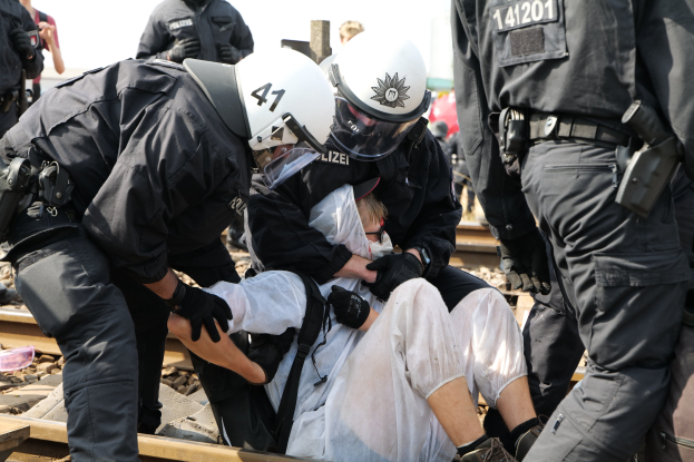 Polizeibeamte in Einsatzausrüstung verhaften einen Mann in einem weißen Kleid auf einem Bahnsteig, mit anderen Personen im Hintergrund.