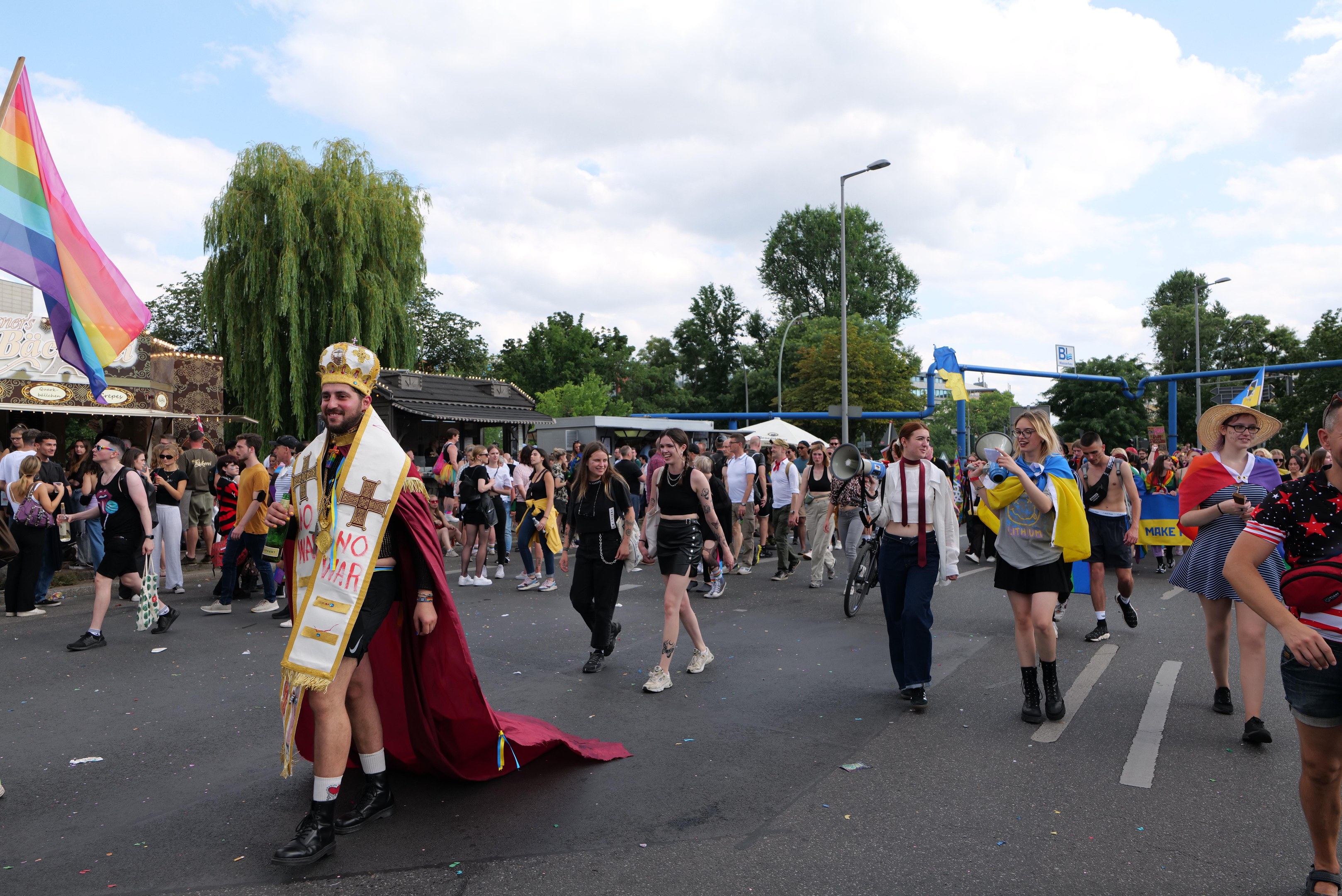 Eine Gruppe von Menschen marschiert bei der Pride Parade 2018 mit einer Regenbogenflagge und Musikinstrumenten, einige tragen Mützen, vor einem Hintergrund aus Laternenmasten, Bäumen, Schuppen und einem bewölkten Himmel.