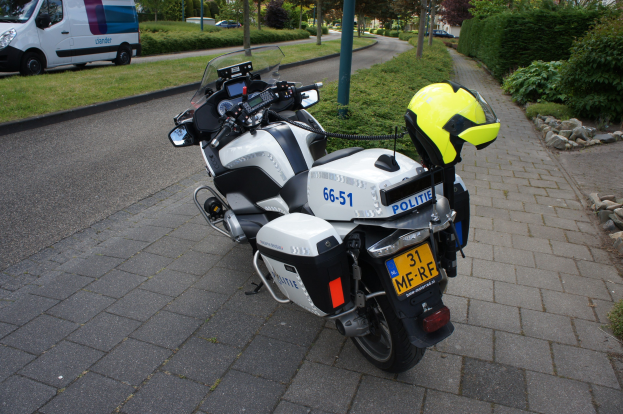 Ein Polizeimotorrad am Straßenrand geparkt mit einem Helm darauf, umgeben von Bäumen, Gebäuden und anderen Fahrzeugen.