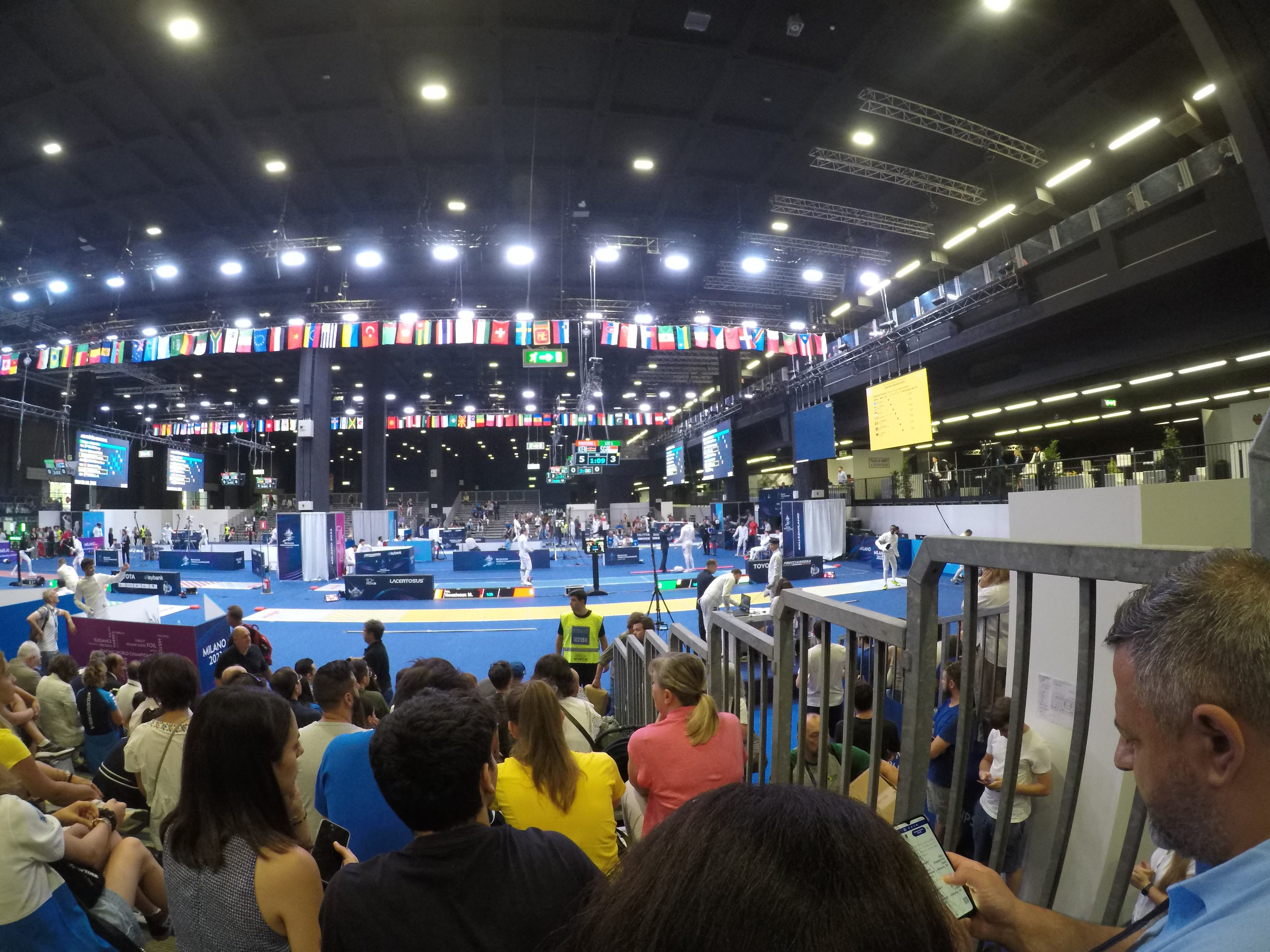 Große Menschenmenge sitzt und steht in einer Indoor-Sportarena mit Bildschirmen, Flaggen und Deckenbeleuchtung im Hintergrund.
