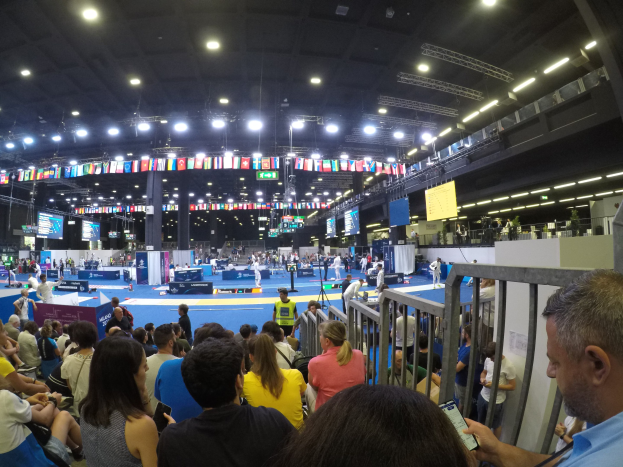 Große Menschenmenge sitzt und steht in einer Indoor-Sportarena mit Bildschirmen, Flaggen und Deckenbeleuchtung im Hintergrund.