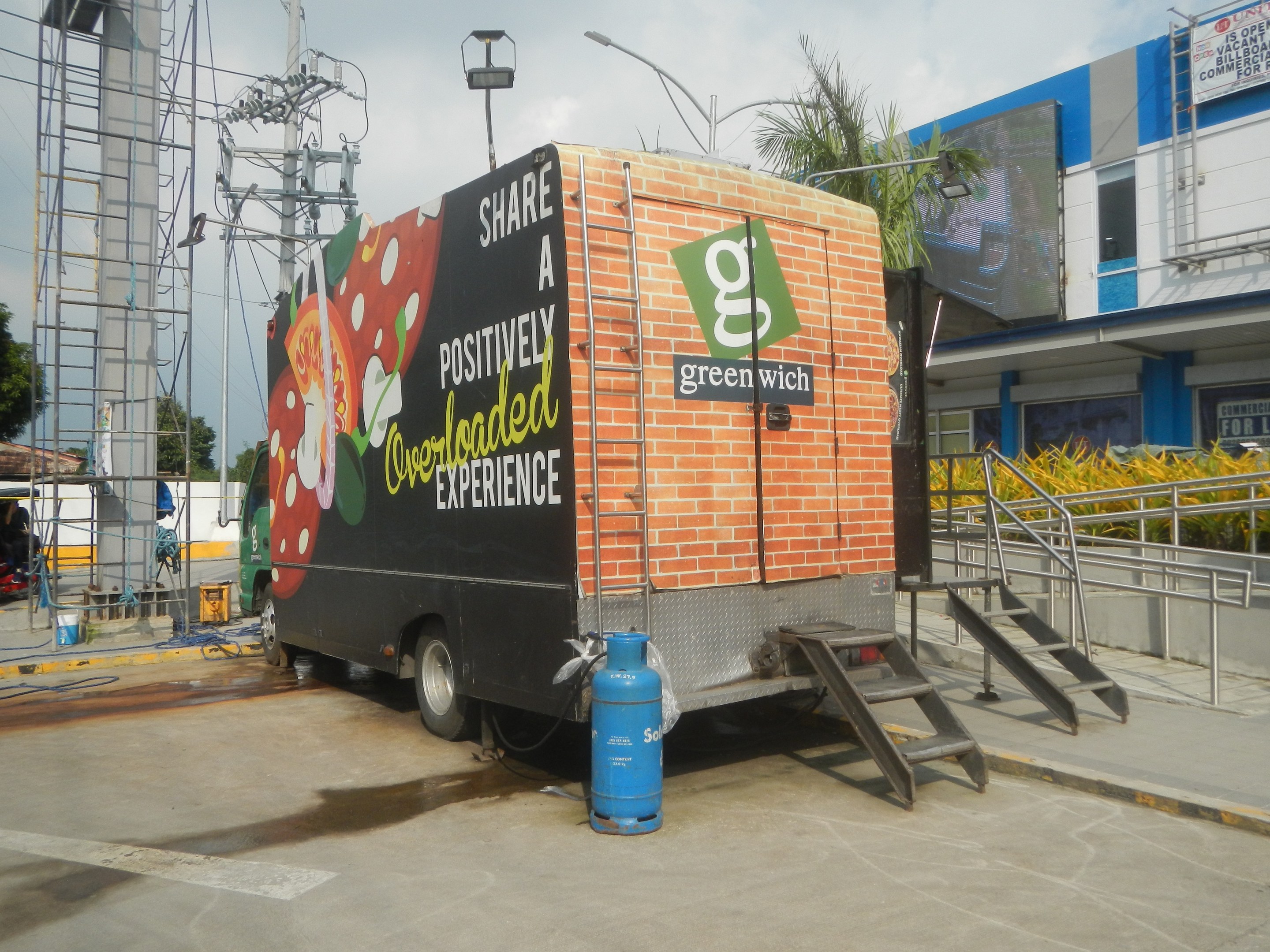 Ein großer Lastwagen parkt vor einem Gebäude mit einer Tafel, auf der "Teilen Sie eine positiv überladene Erfahrung" steht, mit Stufen, einem Zylinder auf dem Boden, Geländern, Strommasten, Laternen, Bäumen und einem bewölkten Himmel im Hintergrund.