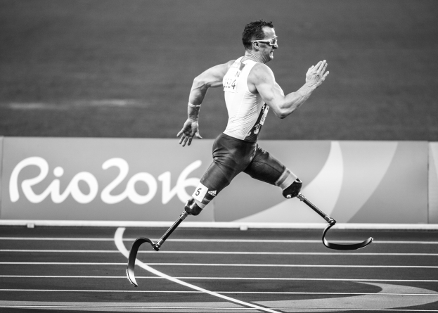 Schwarzes und weißes Foto eines Mannes mit einem Prothesenbein, der auf einer Laufbahn läuft, mit einer Tafel im Hintergrund, auf der der Text 'Rio 2016 Paralympic Games' angezeigt wird.
