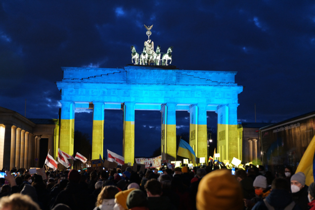 Menge mit Fahnen und Plakaten vor dem Brandenburger Tor, mit einem Banner auf der rechten Seite mit protestbezogenem Text.