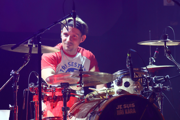 Ein Mann in einem roten und weißen T-Shirt, der auf der Bühne Schlagzeug spielt und vor mehreren Mikrofonen strahlt.