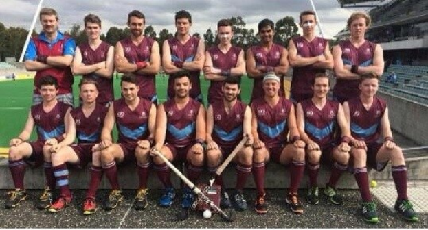 Eine Gruppe von Männern, die auf einem Feld mit Hockey-Schlägern feiern, Bäume und ein Stadion mit Zuschauern im Hintergrund.