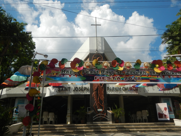Ein Gebäude mit einem Schild, auf dem "Frohes Fest St. Joseph der Arbeiter" steht, vor dem es eine Treppe zum Eingang mit einem Bogen und Dekorationen gibt, umgeben von Stühlen, Tischen, Topfpflanzen und Bäumen unter einem bewölkten Himmel.