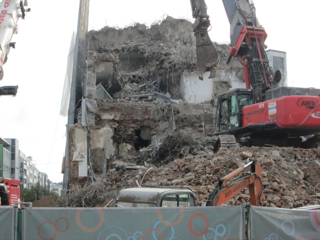 Ein Gebäude wird mit einem Kran abgerissen, umgeben von Kränen, Fahrzeugen, Felsen und einer Fahne mit Text, mit Gebäuden, Bäumen und Himmel im Hintergrund.