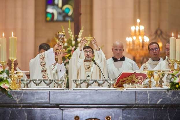 Eine Gruppe von Priestern steht vor einem Altar, jeder hält einen Kelch in der Hand, mit verschiedenen Gegenständen auf einem zentralen Tisch, einem Blumenstrauß links, einem Kerzenständer rechts und einem Bleiglasfenster im Hintergrund.