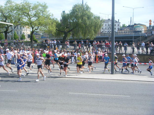 Gruppe von Menschen beim Marathon auf einer Straße mit Absperrungen, Zuschauern und einem Ziellinienband unter einem bewölkten Himmel.