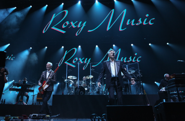 Eine Gruppe von Musikern, die auf der Bühne mit Mikrofonen und Ständern spielen, Kabel im Vordergrund und ein Hintergrund mit der Aufschrift "Roxy Music - Live at the O2 Arena, London."