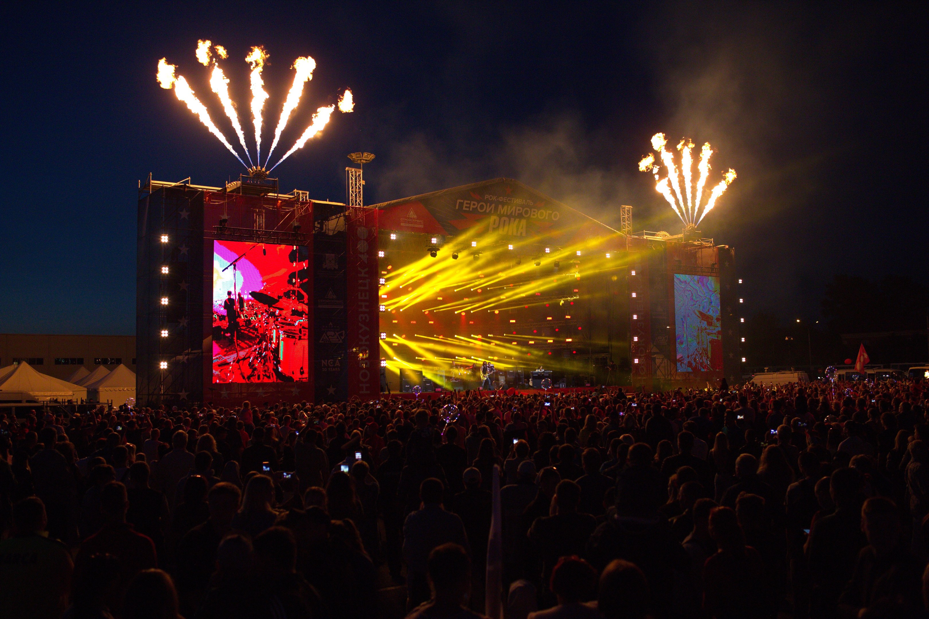 Große Menschenmenge auf einem Musikfestival mit beleuchteter Bühne, die Musiker, Banner, Zelte links und Feuerwerk am dunklen Himmel zeigt.
