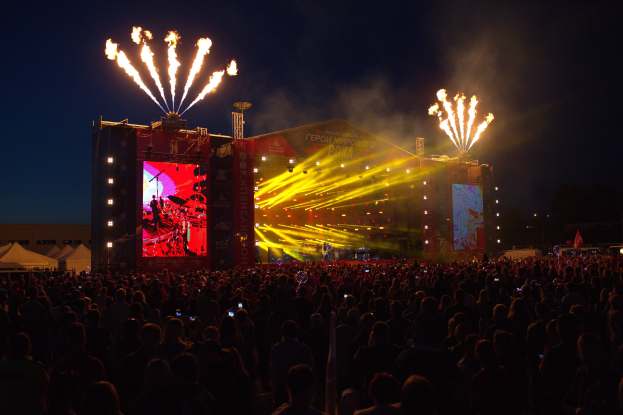 Große Menschenmenge auf einem Musikfestival mit beleuchteter Bühne, die Musiker, Banner, Zelte links und Feuerwerk am dunklen Himmel zeigt.