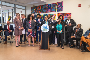 Eine Gruppe von Menschen steht vor einem Rednerpult mit Mikrofon und einer Tafel mit Text, während andere auf Stühlen sitzen oder in der Nähe stehen; Flaggen, ein Feuerlöscher und Deckenleuchten sind an der Rückwand zu sehen.