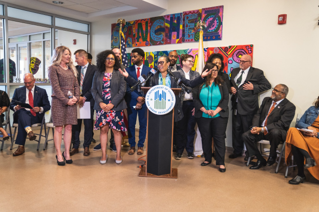 Eine Gruppe von Menschen steht vor einem Rednerpult mit Mikrofon und einer Tafel mit Text, während andere auf Stühlen sitzen oder in der Nähe stehen; Flaggen, ein Feuerlöscher und Deckenleuchten sind an der Rückwand zu sehen.
