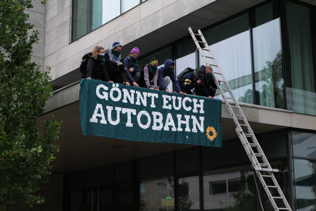 Gruppe von Menschen auf einem Gebäude, die eine Fahne halten, mit einem Baum links und Glasfenstern im Hintergrund, die scheinbar protestieren und die deutsche Regierung unterstützen.