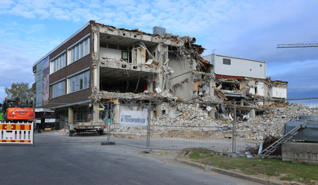Ein großes abgerissenes Gebäude in der Mitte einer Straße, umgeben von einem Metallzaun, mit Fahrzeugen, Gras, einem Schild, einer Fahne, Pfählen, einem Kran, Bäumen und einem bewölkten Himmel.