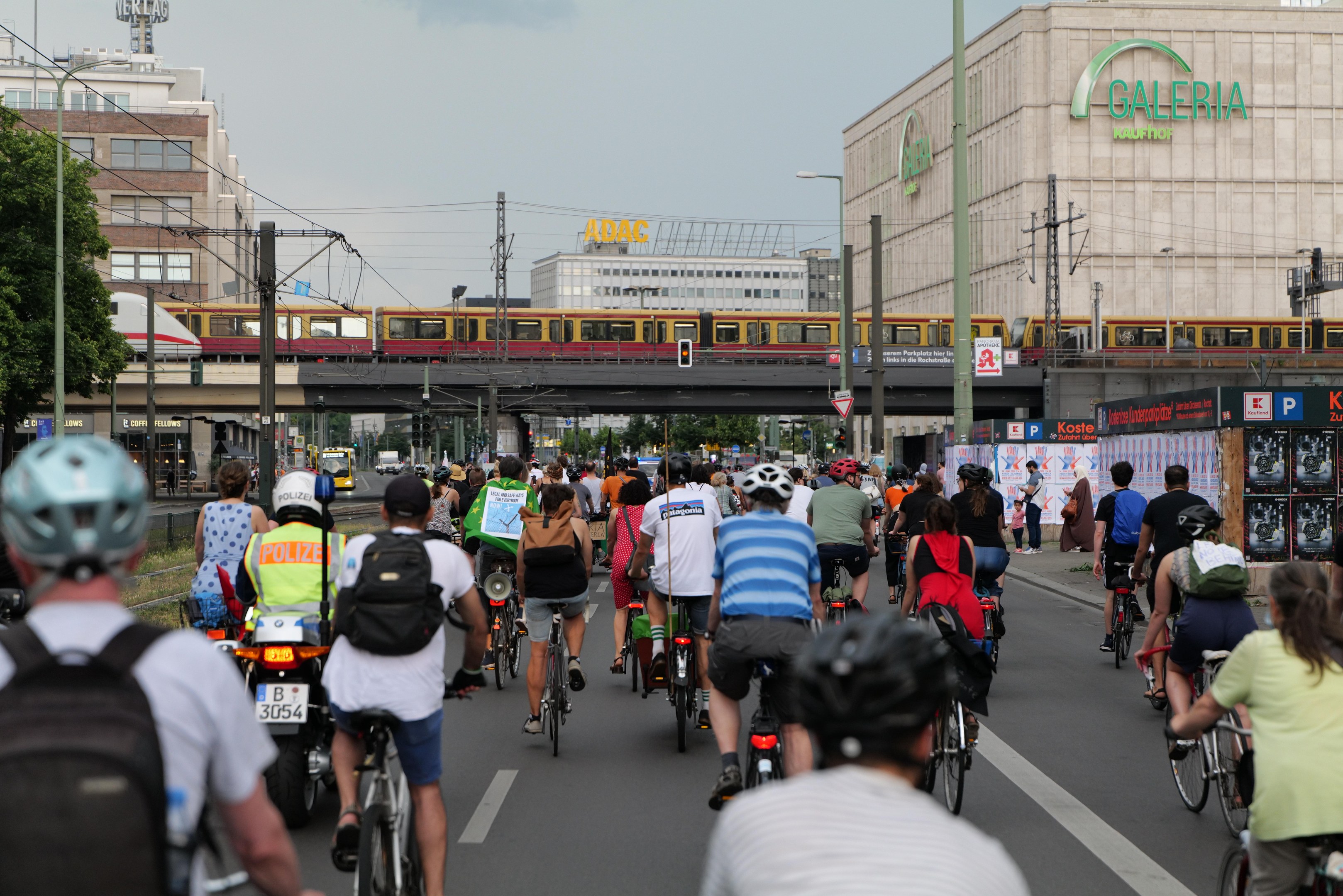 Eine Gruppe von Menschen, die auf Fahrrädern eine Straße mit hohen Gebäuden entlangfahren, einige tragen Helme und Taschen, mit einem Zug auf einem Bahngleis, Strommasten, Bäumen und einem klaren blauen Himmel im Hintergrund.