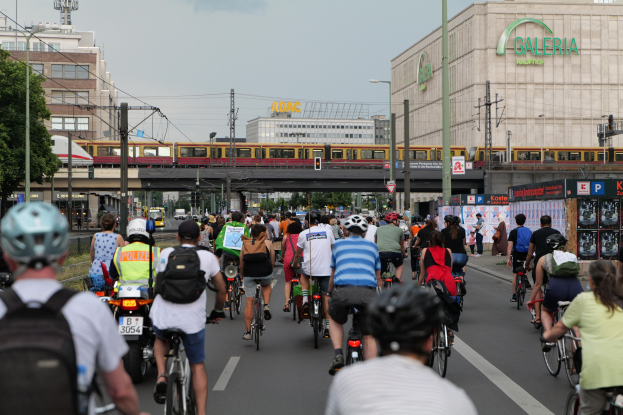 Eine Gruppe von Menschen, die auf Fahrrädern eine Straße mit hohen Gebäuden entlangfahren, einige tragen Helme und Taschen, mit einem Zug auf einem Bahngleis, Strommasten, Bäumen und einem klaren blauen Himmel im Hintergrund.
