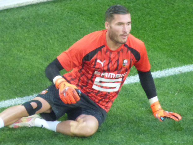 Ein Fußballspieler in rotem T-Shirt, schwarzen Shorts, weißen Socken und orangen Baseballhandschuhen sitzt auf dem Boden mit Händen auf den Knien.