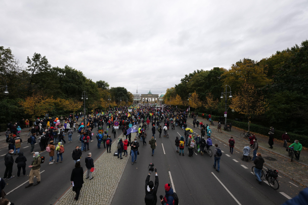 Eine große Gruppe von Menschen marschiert eine von Bäumen gesäumte Straße in Berlin entlang und hält Kameras während einer Protestaktion, mit einem Gebäude und einem klaren Himmel im Hintergrund.