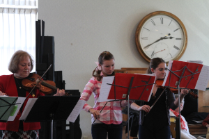 Drei Frauen auf der Bühne mit Violinen, Musikständern und Notenblättern, während ein Zuschauer im Vordergrund und eine Uhr an der weißen Wand im Hintergrund zu sehen ist, mit einem weißen Vorhang in der linken Ecke.