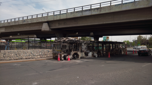 Ein ausgebrannter Bus steht am Straßenrand, umgeben von Absperrbaken, Steinen, einem Zaun, Bäumen, Straßenlaternen, einem Schild, einer Brücke mit Text, Drähten und unter einem bewölkten Himmel.