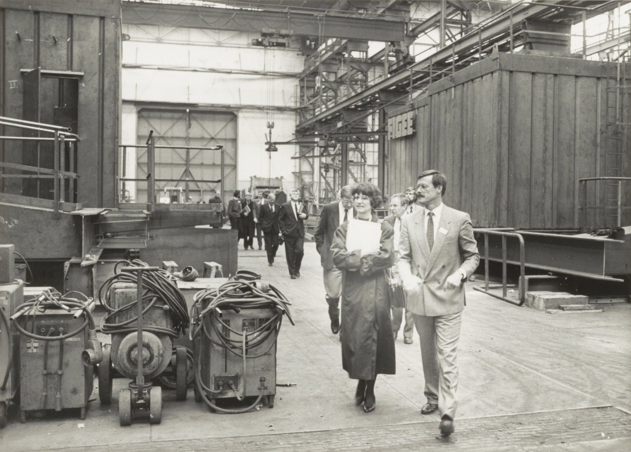 Eine Gruppe von Menschen in Anzügen geht durch eine Fabrik mit Maschinen auf der linken Seite und einer Wand im Hintergrund.