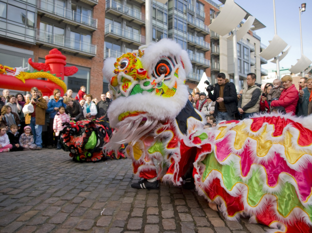 Ein lebendiges chinesisches Neujahrsfest in Amsterdam mit einem Löwen tanzen im Vordergrund und einer Menschenmenge, einige halten Kameras, vor einem Hintergrund von Gebäuden, Laternenmasten und einem klaren blauen Himmel.