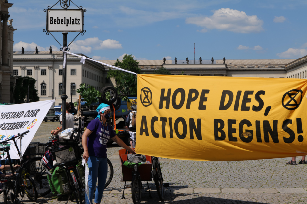 Eine Gruppe von Menschen steht vor einem Gebäude und hält ein gelbes Banner mit der Aufschrift "Hope Dies, Action Begins". In der Nähe stehen Fahrräder und ein Mast mit einer Tafel, unter einem klaren blauen Himmel mit Bäumen im Hintergrund.