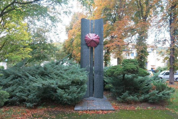 Große Metallskulptur inmitten eines Parks mit Bäumen, Pflanzen, Gras und verstreuten Blättern, umgeben von Gebäuden und einem klaren blauen Himmel, Teil der Kunst der Stadt Berlin.