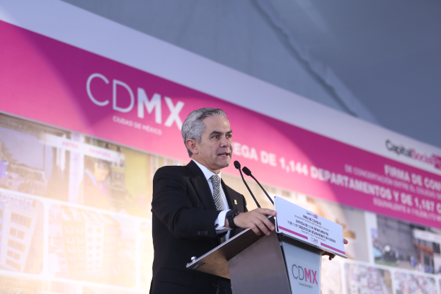 Ein Mann im Anzug und Krawatte steht hinter einem Podium mit zwei Mikrofonen und einer Tafel, vor ihm eine Fahne mit Bildern und Text.