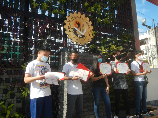 Gruppe von Menschen in Masken, die Zertifikate halten und vor einem Universitätsgebäude mit einer Pflanzenwand links und einem Bogen mit Logo rechts stehen.