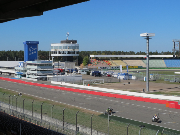Eine Gruppe von Menschen, die Motorräder auf einer Rennstrecke fahren, umgeben von einem Metallzaun, Gras und einem bewölkten Himmel.