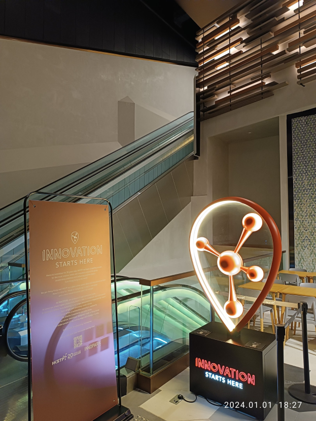Großes Schild in einem Gebäude mit einer Rolltreppe im Hintergrund, das Tische und Stühle auf der rechten Seite und eine Tafel mit 'Innovation beginnt hier' auf der linken Seite zeigt, was auf die HSTP20 Expo hinweist.
