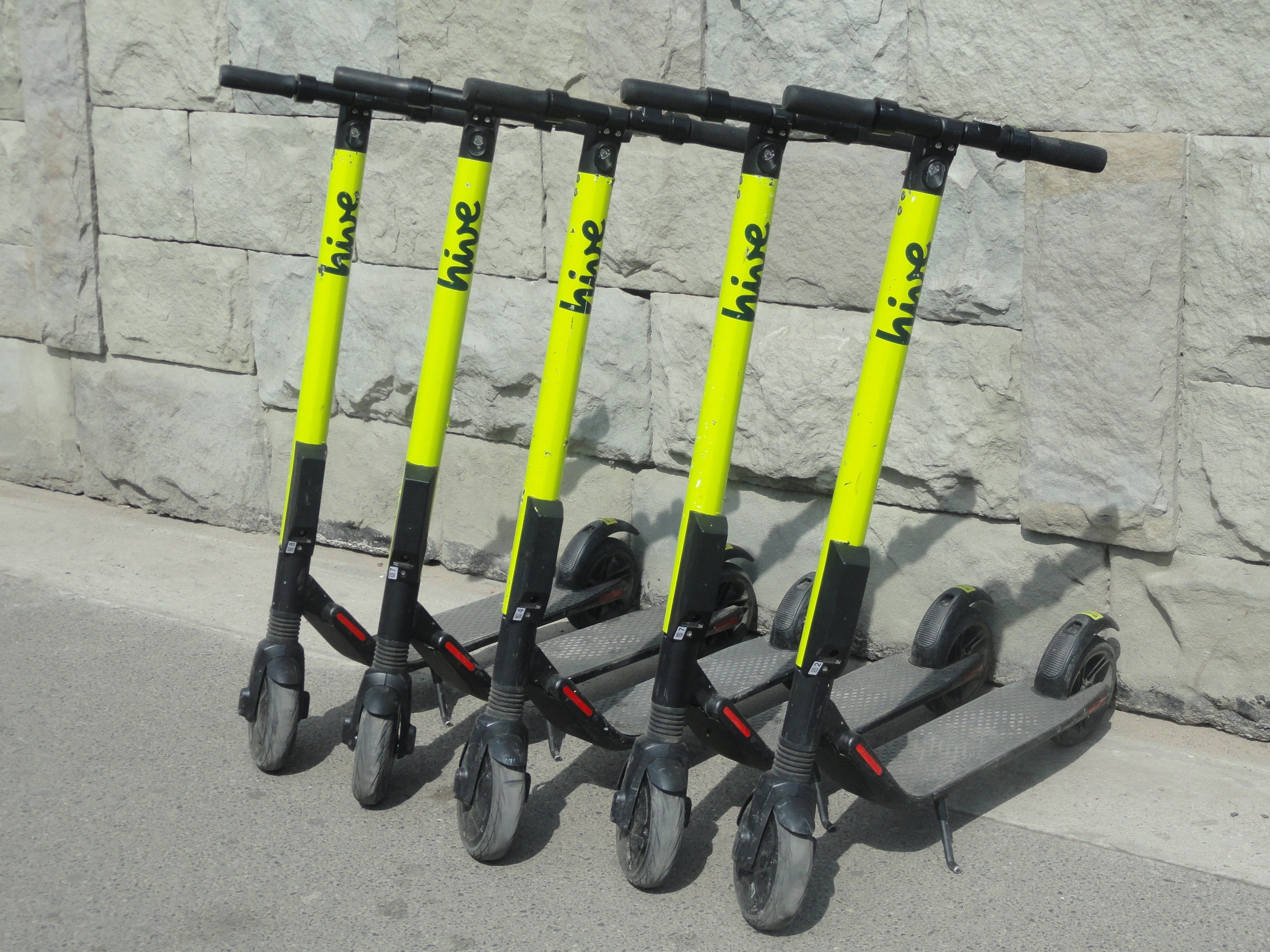 Drei schwarze und gelbe E-Scooter nebeneinander auf einem Straßenrand mit einer Wand im Hintergrund geparkt.