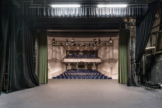 Großes Auditorium mit schwarzen Vorhängen, einer beleuchteten Bühne und Sitzreihen, mit Gegenständen auf der rechten Seite, die auf eine Veranstaltungsstätte hinweisen.