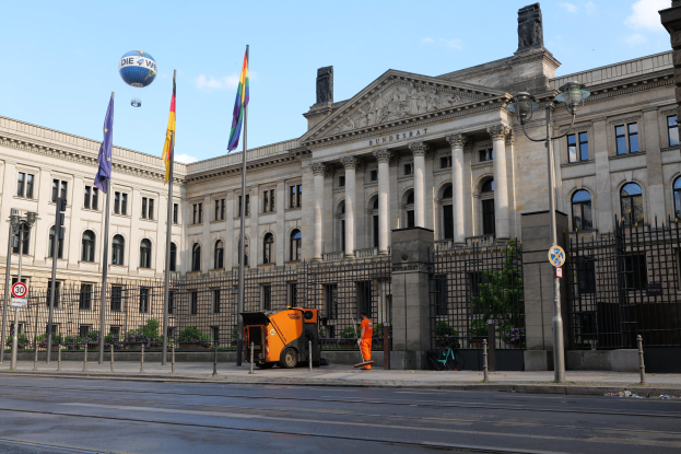 Großes modernes Gebäude mit zahlreichen Fenstern, als Bundestag in Berlin, Deutschland identifiziert, umgeben von Straßeninfrastruktur, Fahrzeugen, Passanten, Flaggen und Bäumen unter einem bewölkten Himmel.
