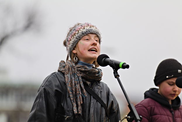 Eine Frau in einer schwarzen Jacke, einem Schal und einer Mütze singt in ein Mikrofon auf der Bühne, während eine andere Person in einer maronenfarbenen Jacke und einer schwarzen Mütze neben ihr steht, und eine unscharfe Menge zusieht.