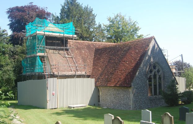 Kirche mit einer grünen Plane auf dem Dach, umgeben von einem Metallzaun, Bäumen, Pflanzen, Gras, einem Weg, Pfosten und einem bewölkten Himmel.